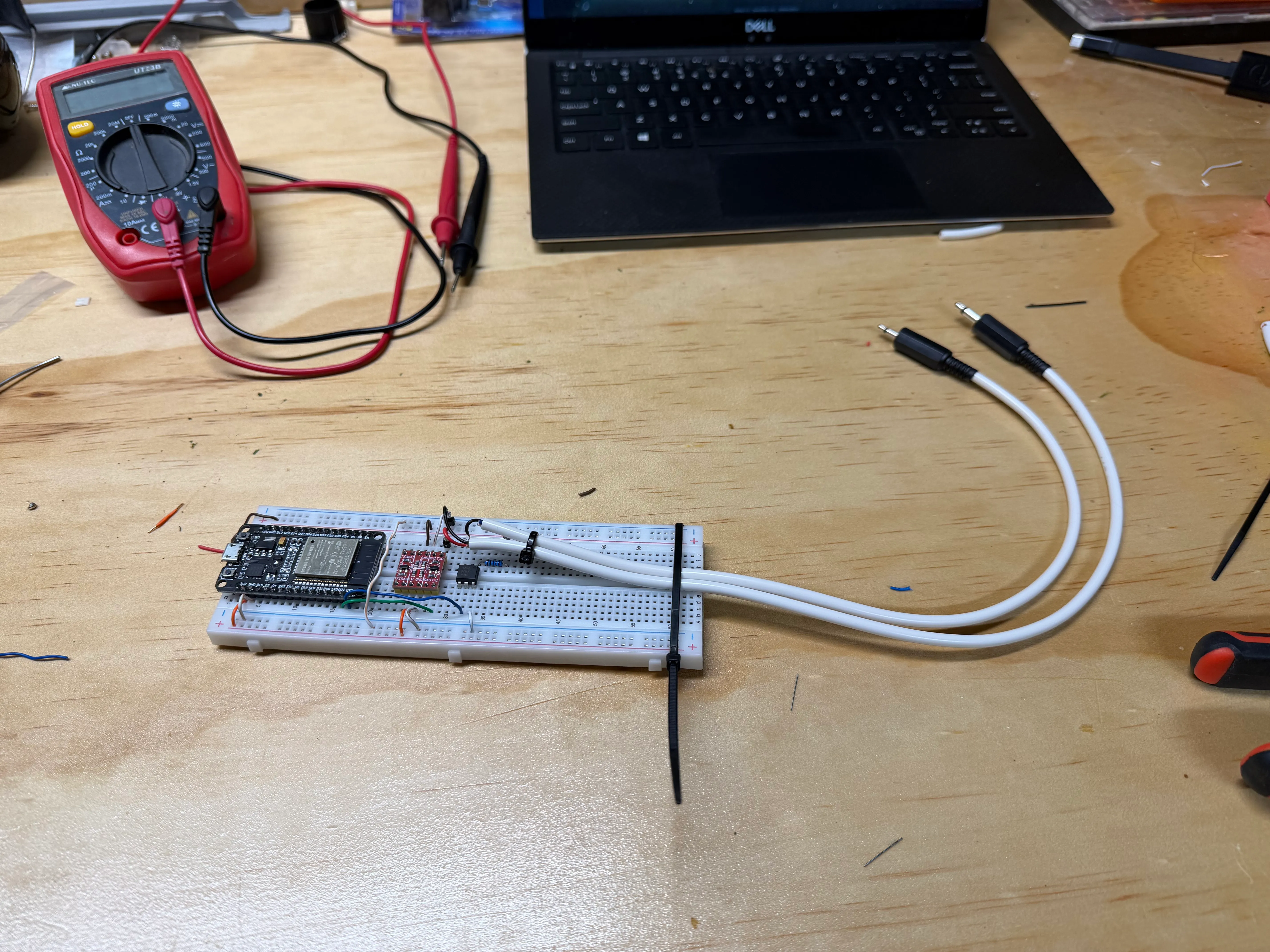 A prototyping breadboard on a workbench with 3 electronic components: a micro controller, a logic level shifter and an optocoupler. There are jumper wires connecting the components together and two grey leads with black audio plugs extending away from the board. There is a red multimeter and a laptop in the background.